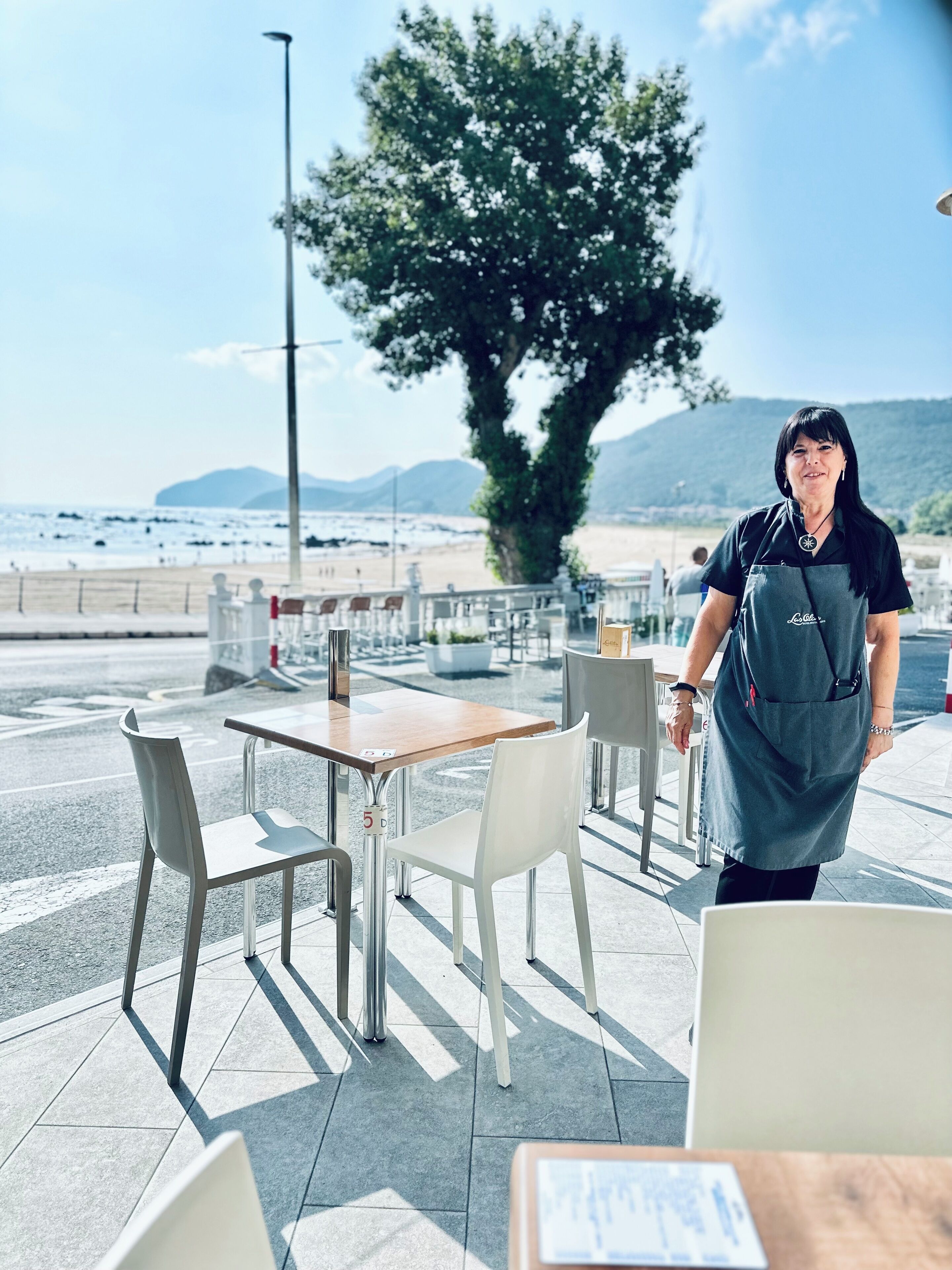 Una persona con uniforme de pie cerca de una mesa y sillas en un patio al aire libre, con vista a la playa y las montañas al fondo.