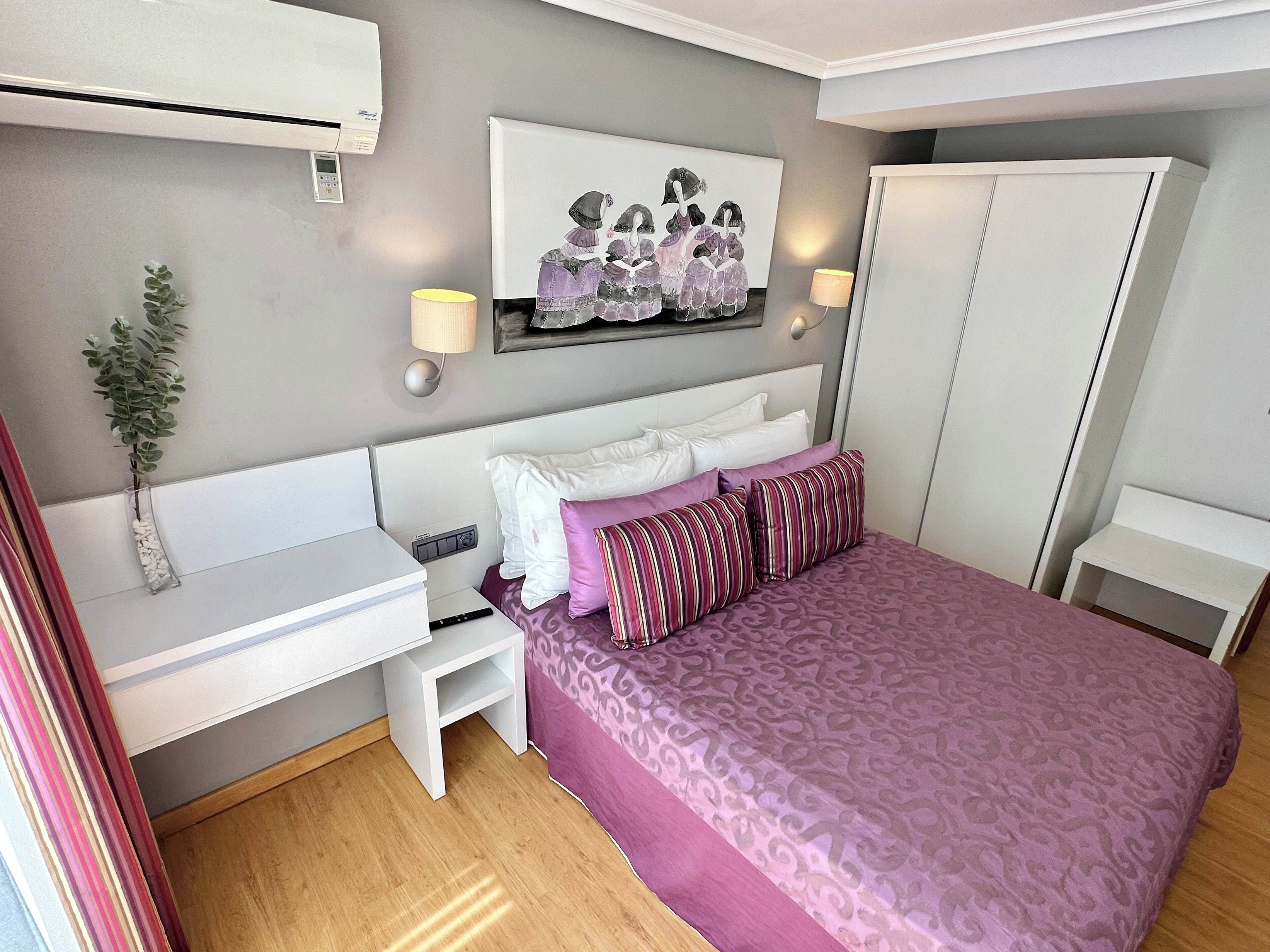 A bedroom with a bed, a desk, a chair, a wardrobe, and a wall-mounted air conditioner.