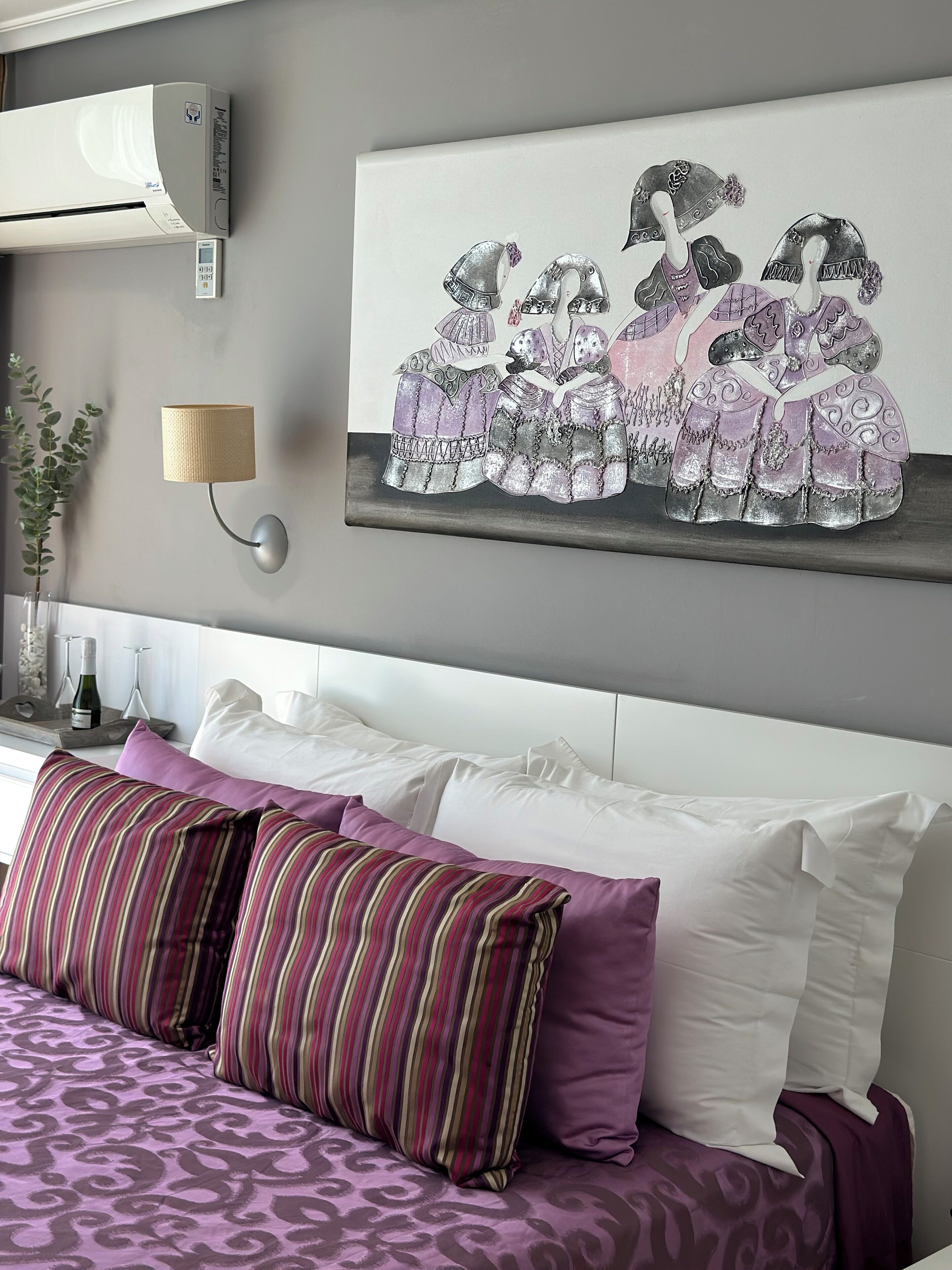 Een hotelkamer met een bed, kussens, een wandairco, een wandlamp en een schilderij van vrouwen in traditionele kleding.