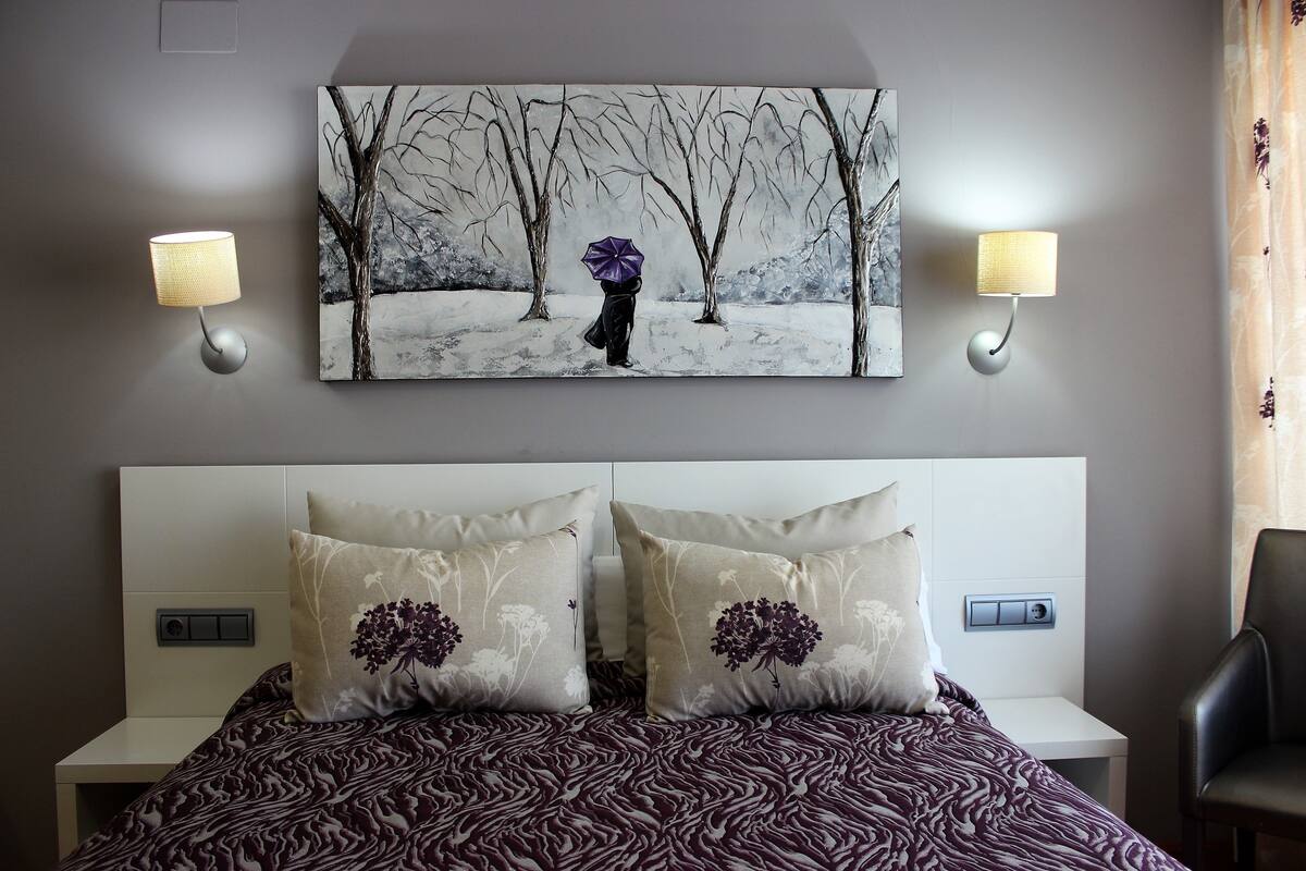 A bedroom with a bed, two wall-mounted lamps, a painting, and a nightstand with a switch.