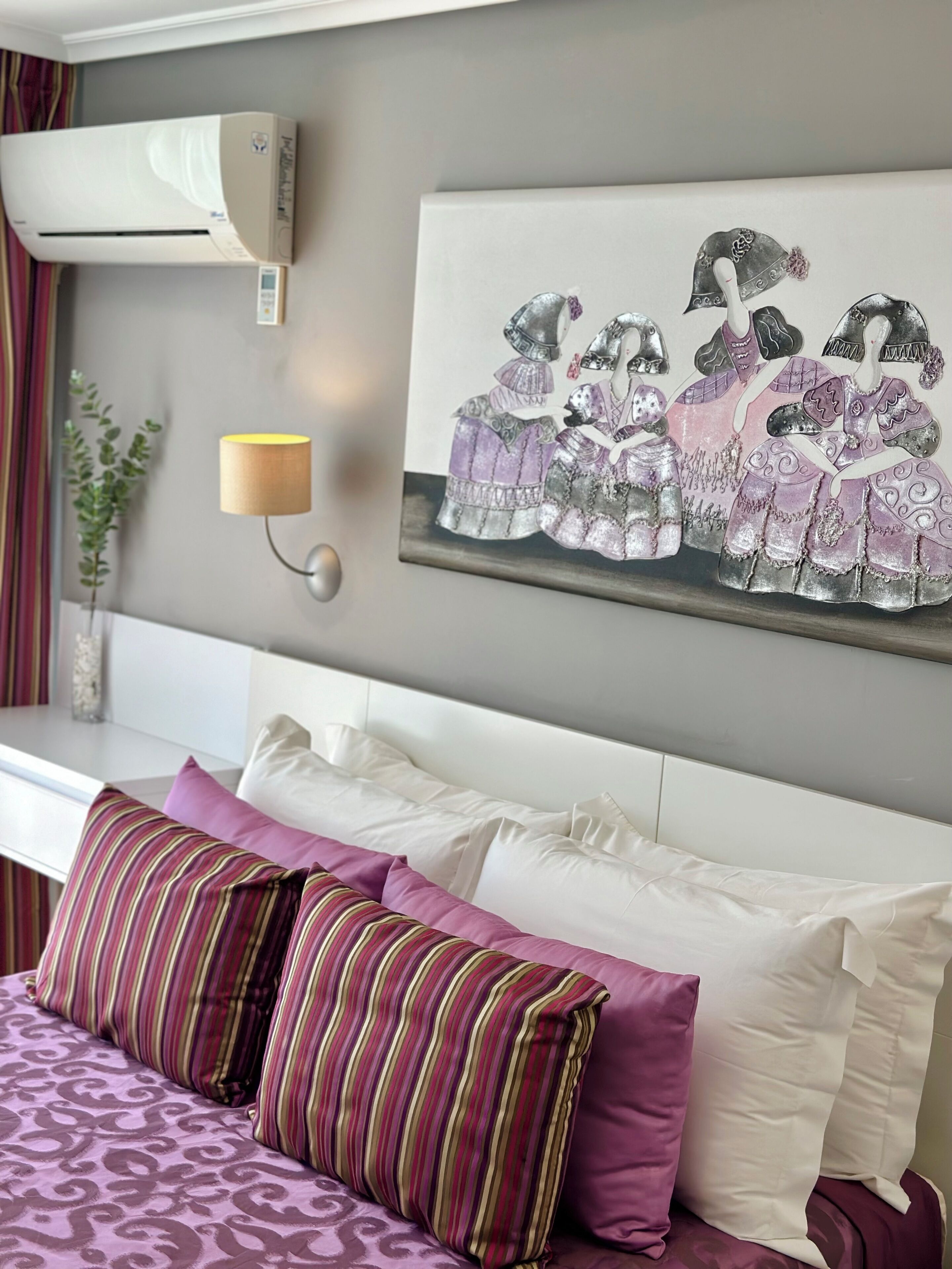 A hotel room with a bed, pillows, a wall-mounted air conditioner, a wall lamp, and a painting of dancers.