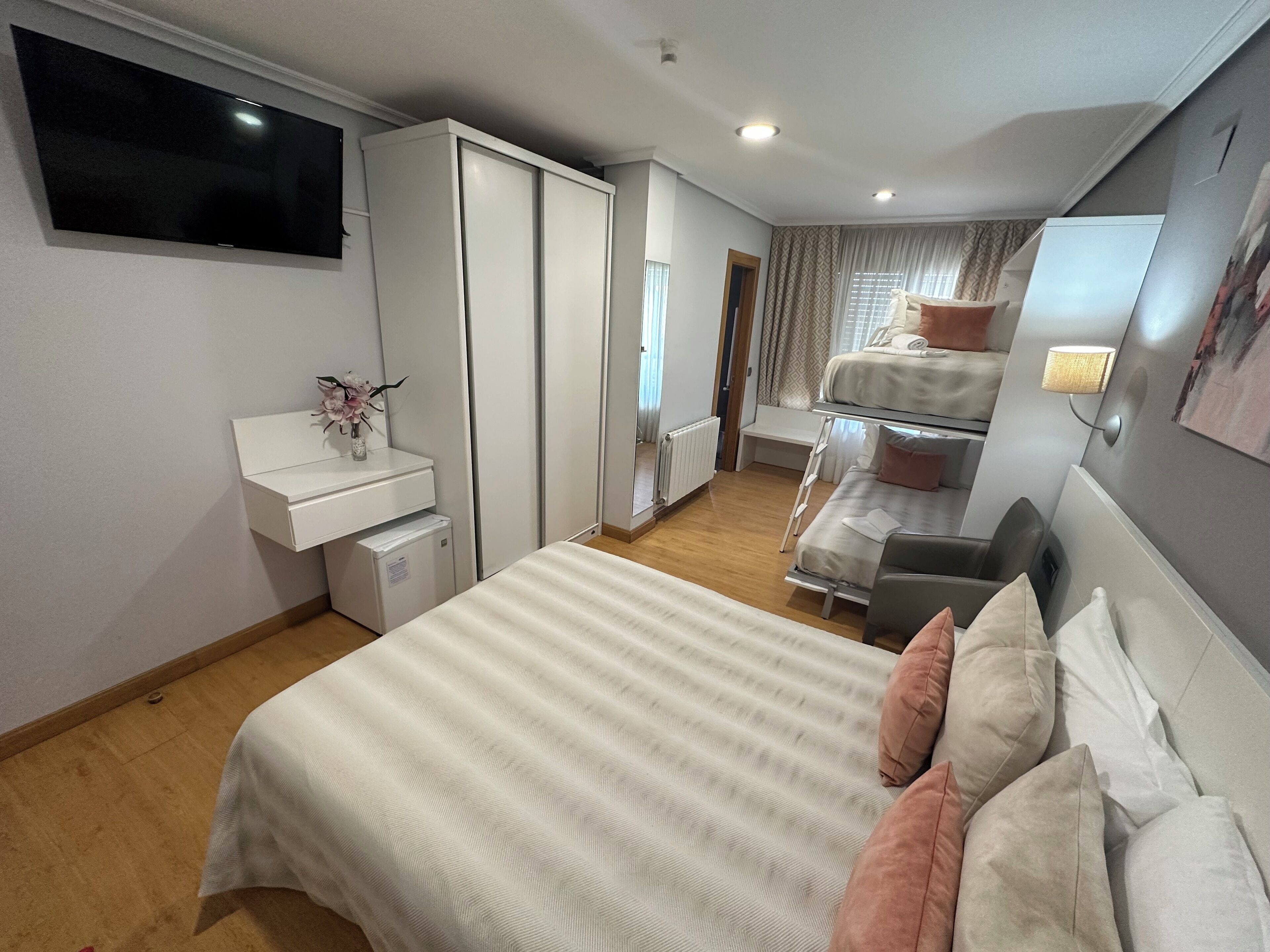 A modern bedroom with a large bed, a desk with a computer, a wardrobe, and a TV mounted on the wall.