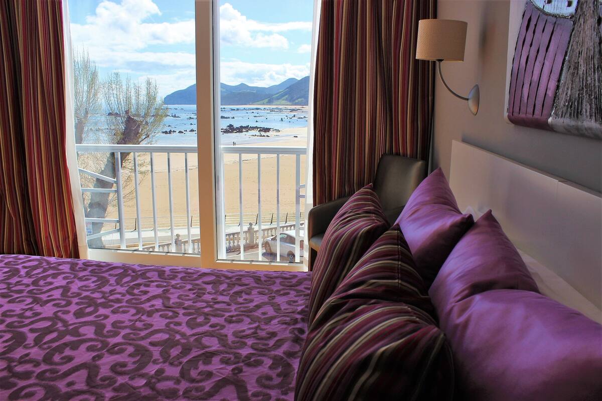A hotel room with a bed, a chair, a view of the beach and mountains, and a lamp.