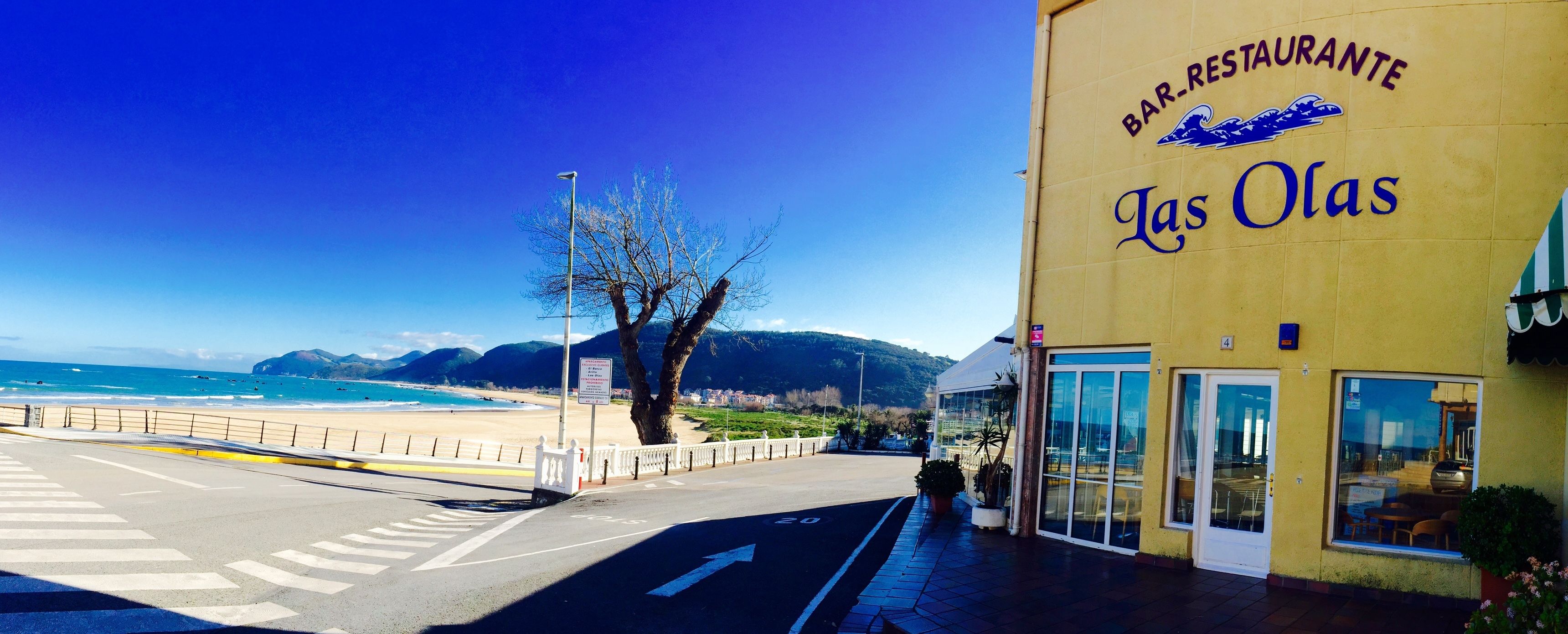 A coastal road with a restaurant named "Las Olas" on the right, featuring a clear view of the beach and mountains in the background.