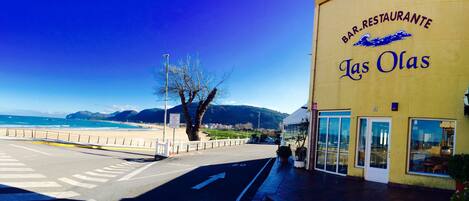 A coastal road with a restaurant named "Las Olas" on the right, featuring a clear view of the beach and mountains in the background.