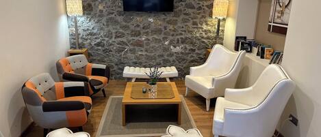 A living room with a stone wall, a flat-screen TV, white leather armchairs, a wooden coffee table, and a bookshelf with books.