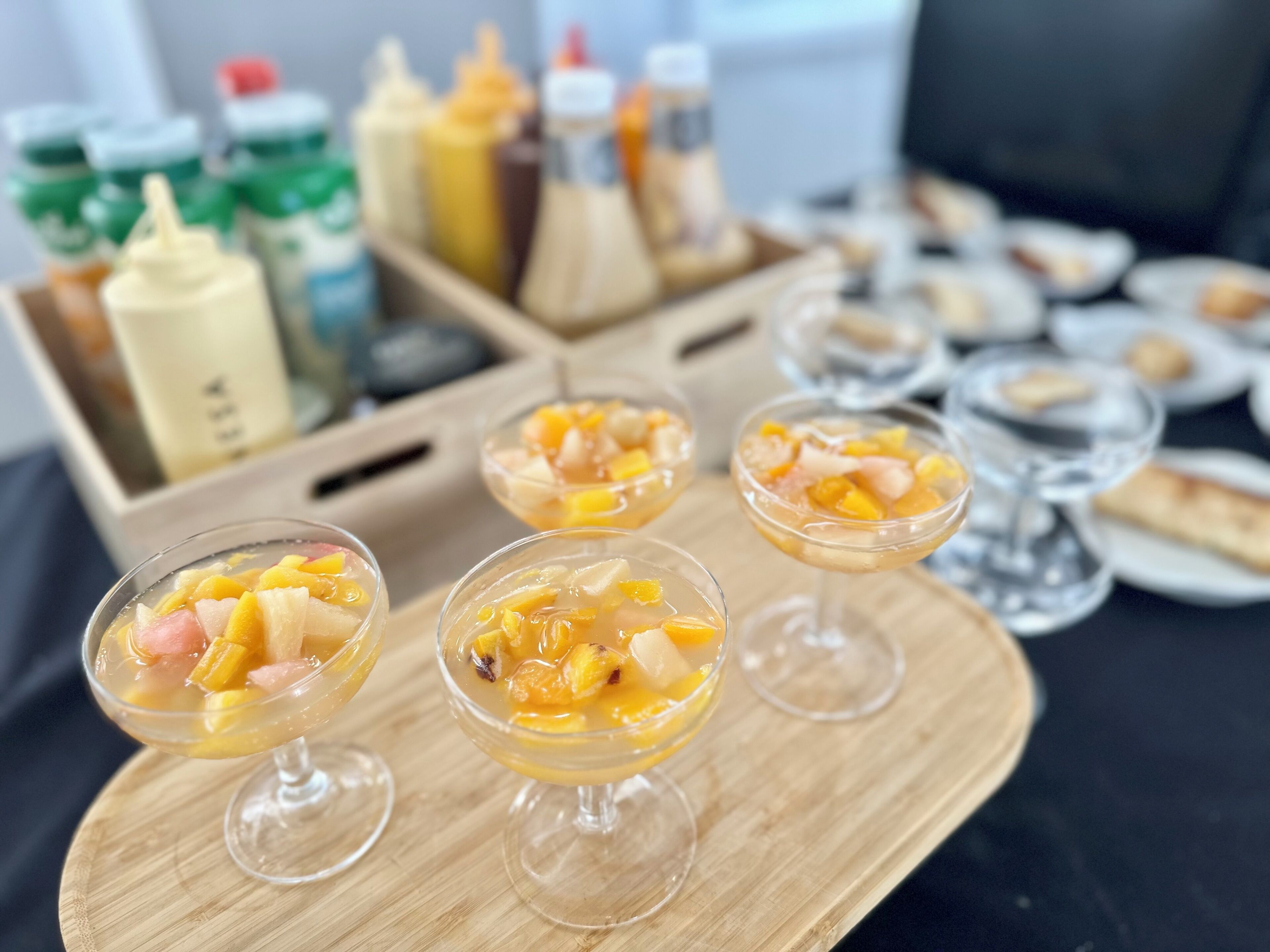 A wooden tray with three glass cups containing a fruit dessert, a condiment station in the background, and plates with food items.