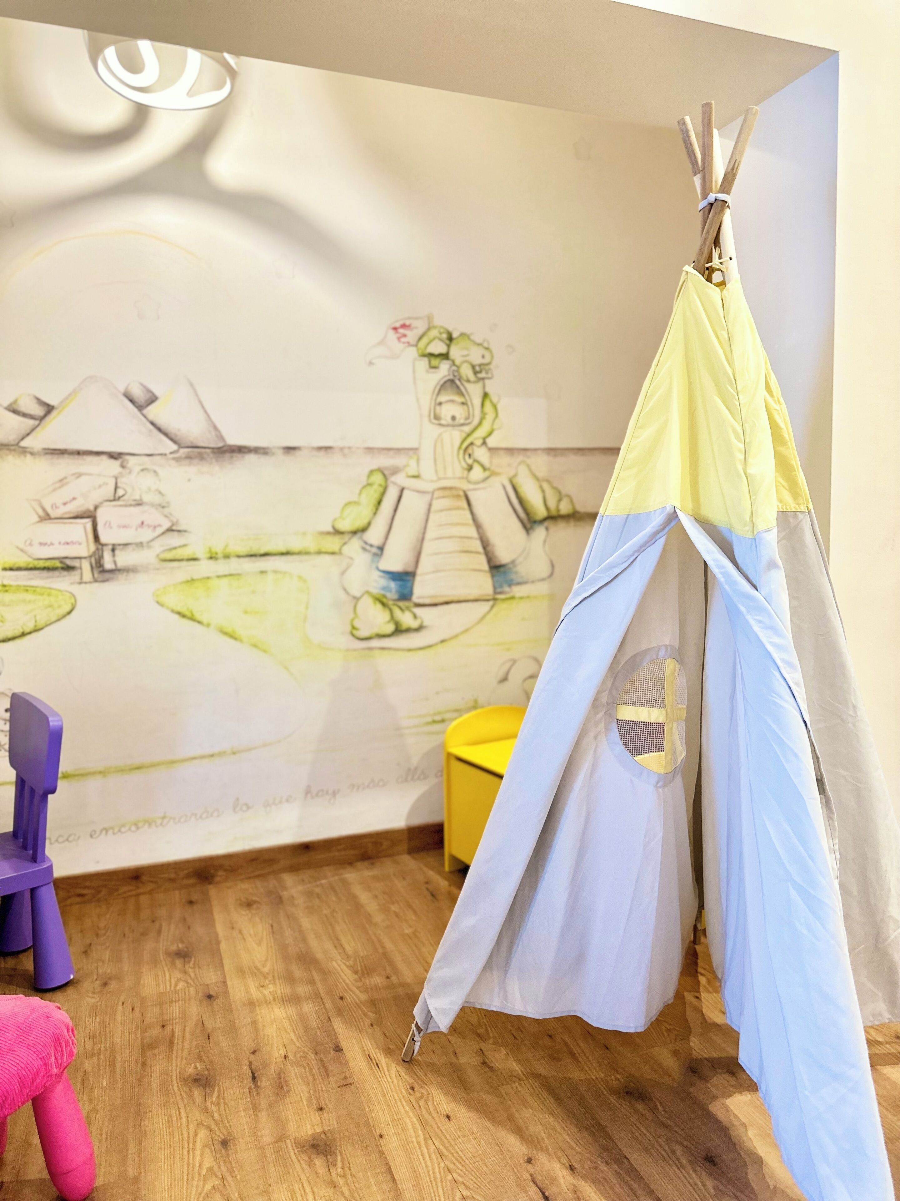 Ein Zimmer mit Holzboden, einem gelben Stuhl, einem weiß-blauen Zelt und einer Wand mit Wandgemälde.