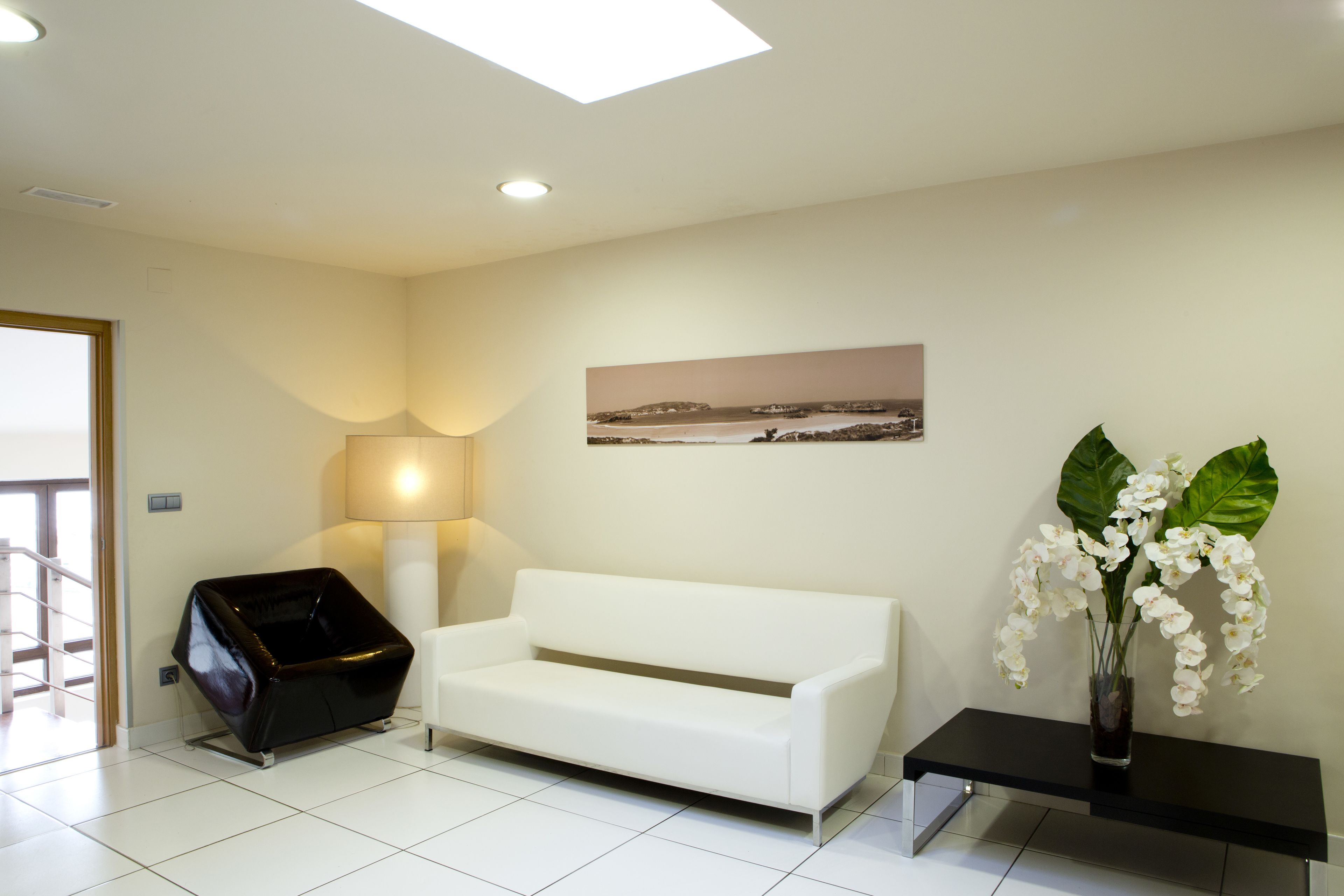 Uma sala de estar moderna com um sofá branco, um banco preto, um abajur e um vaso com flores brancas.