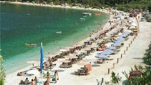 Une plage bordée de rangées de transats et de parasols, un rivage sablonneux et des bateaux sur l’eau.
