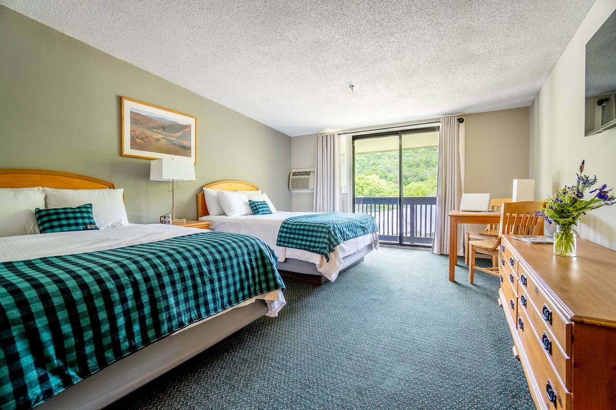 Ein Hotelzimmer mit zwei Betten, einem Schreibtisch, einem Stuhl, einem Laptop und einem Balkon mit Blick.