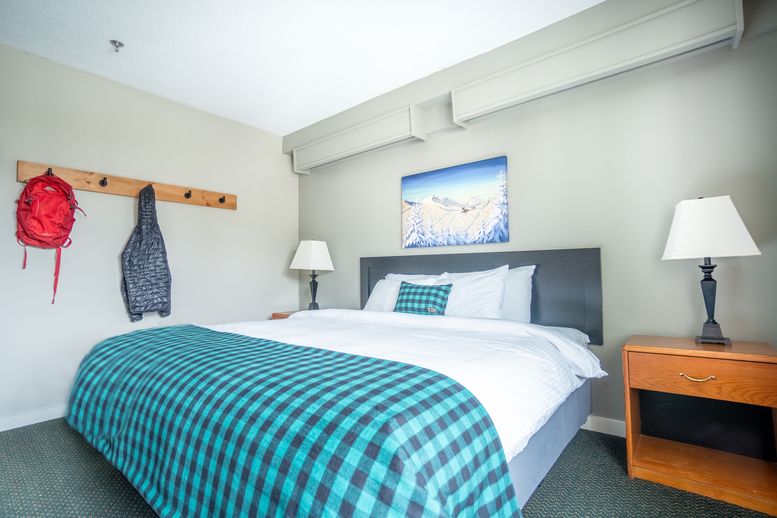 Habitación de hotel con cama, cabecera de madera, mesita de noche, lámpara, perchero con chaquetas y un cuadro en la pared.
