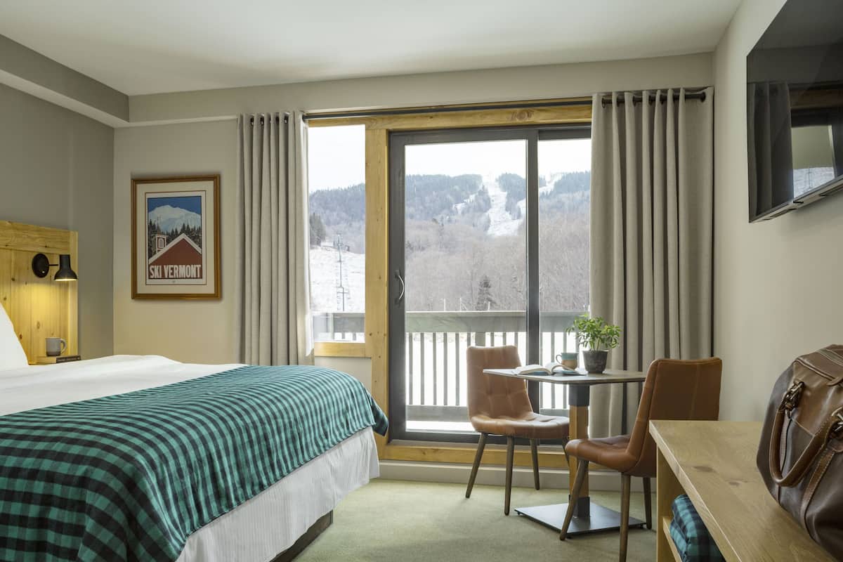 Ein Hotelzimmer mit Bett, Schreibtisch, Stuhl, Fernseher und einem Fenster mit Blick auf Berge, sowie einem gerahmten Bild an der Wand.