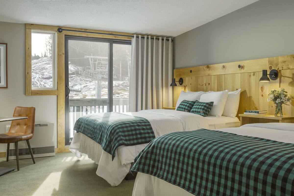 Ein Hotelzimmer mit zwei Betten, einem Holzkopfende, einem Esstisch mit Stühlen und einem großen Fenster mit Vorhängen.