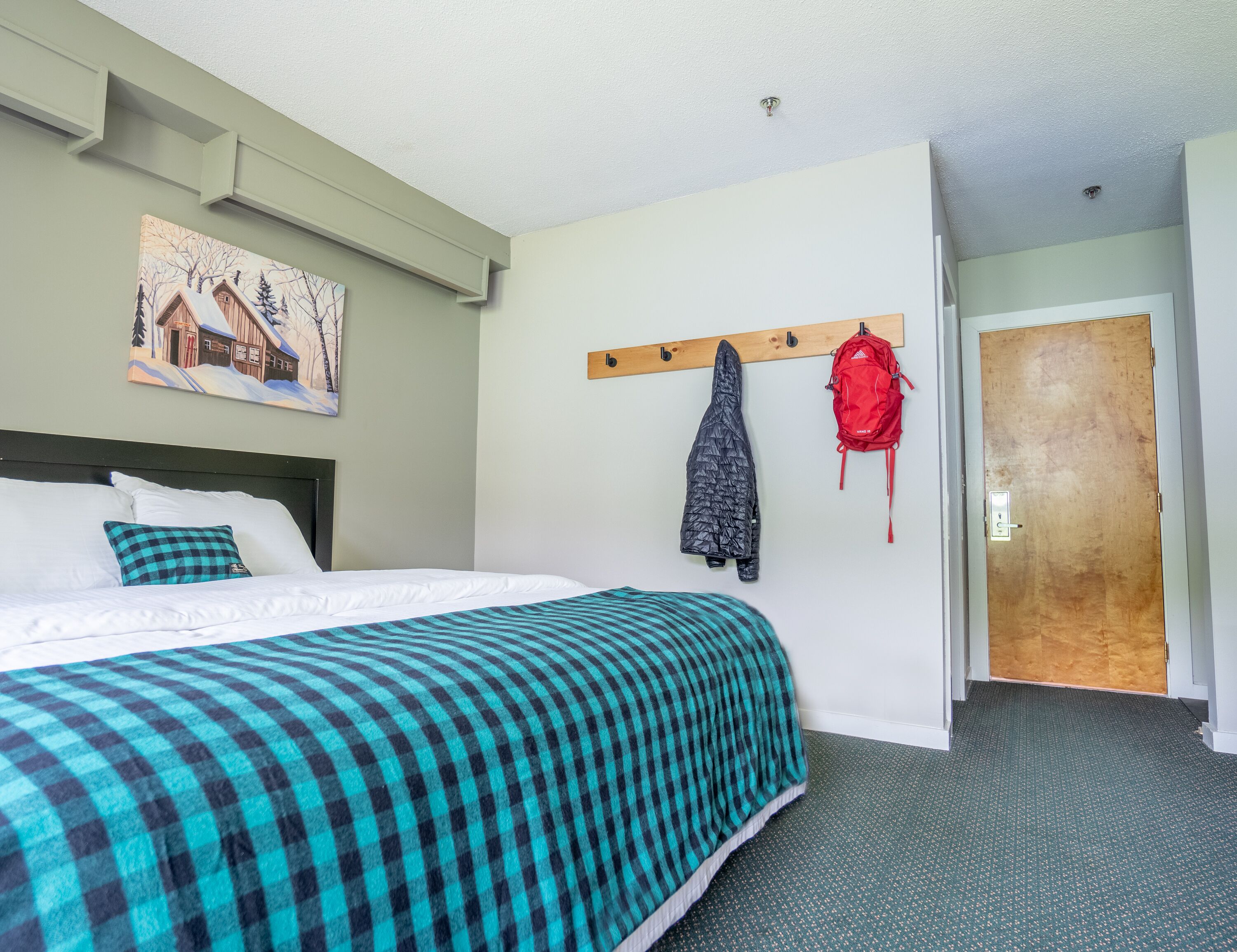 A hotel room with a bed, a coat rack with jackets, a door, and a painting on the wall.