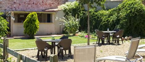 An outdoor patio with tables and chairs, surrounded by a wooden fence, a stone wall, and lush greenery.