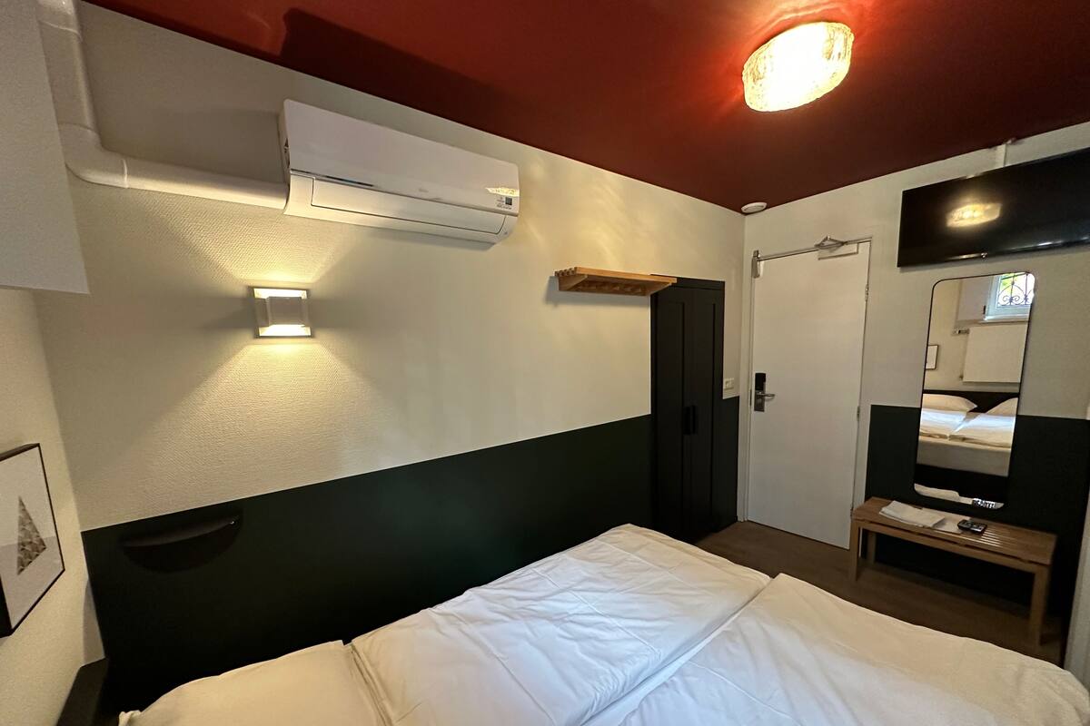 A hotel room with a bed, an air conditioning unit, a bedside table with a lamp, a mirror, and a wardrobe.