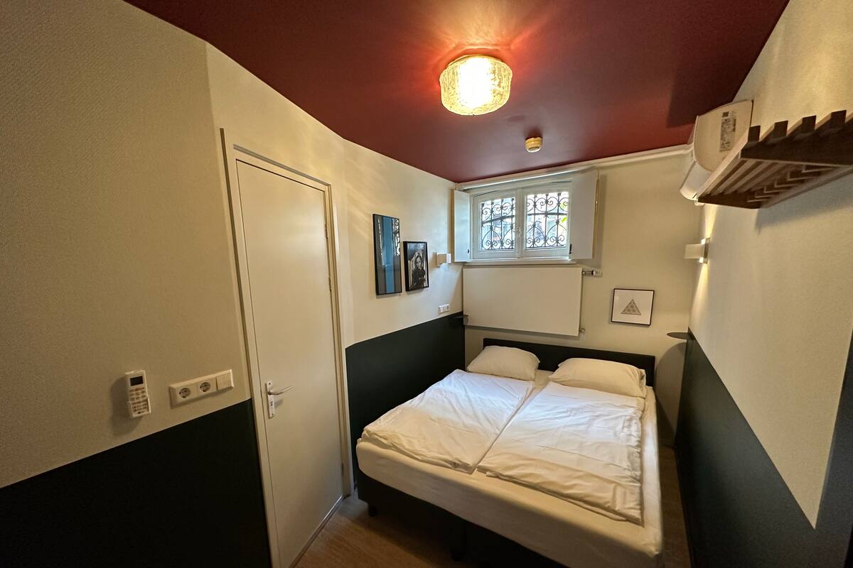 A bedroom with a bed, a window with decorative iron bars, a ceiling light, and a wall-mounted air conditioner.