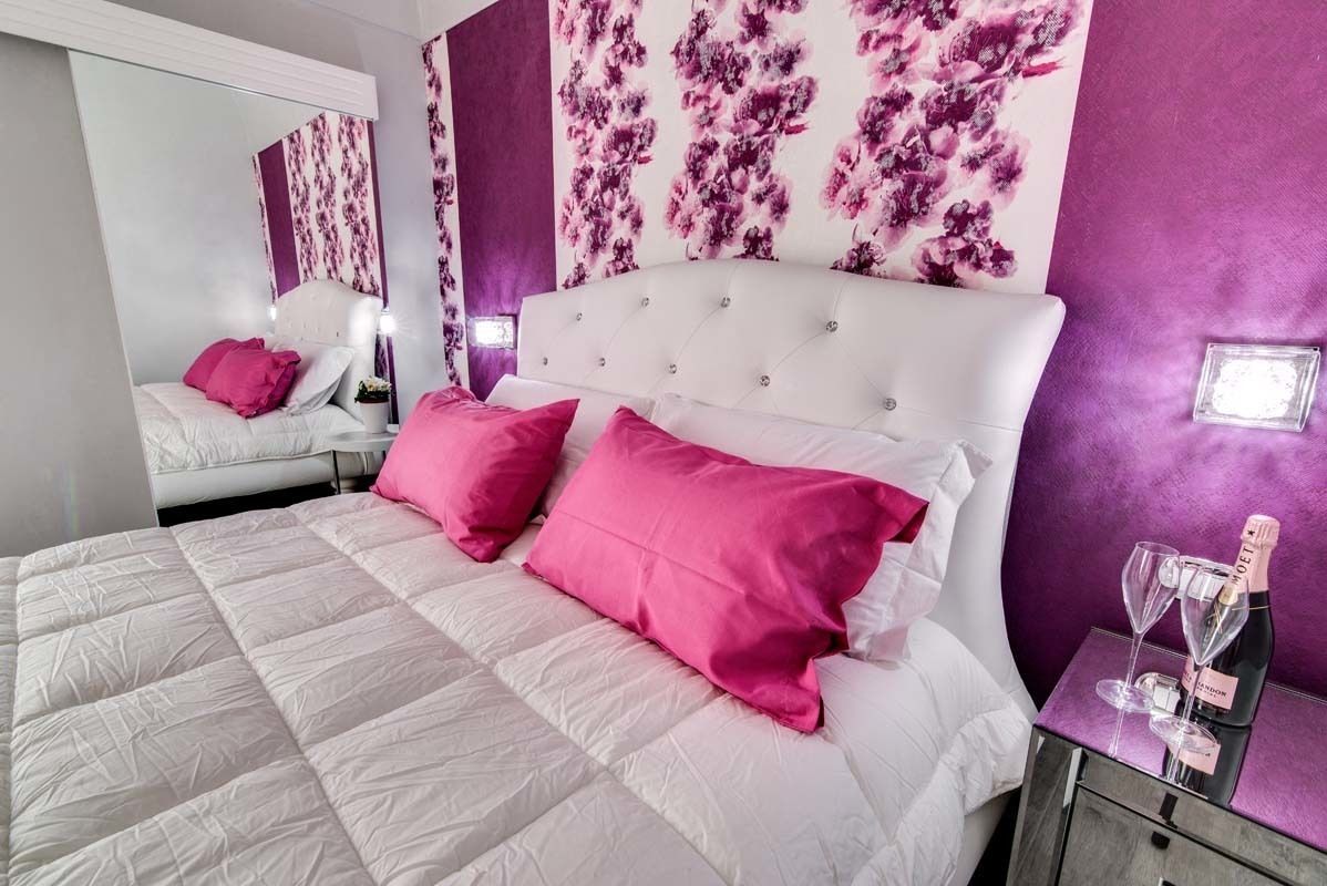A bedroom with a white bed, two pink pillows, a white headboard, and a purple floral wall.