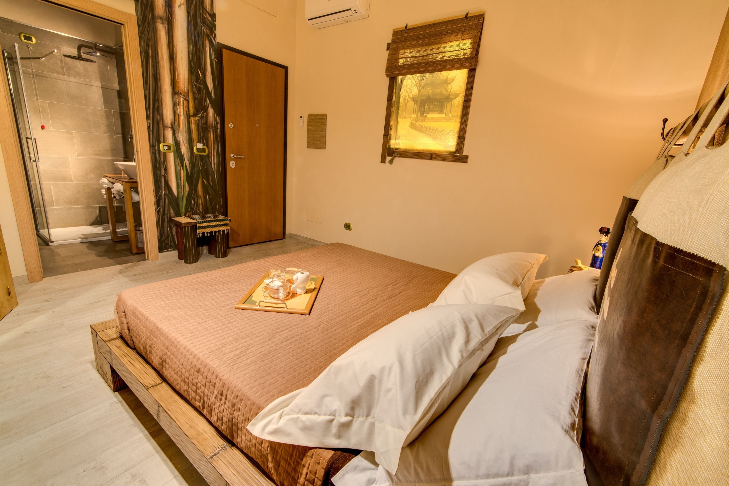 A hotel room with a bed, a wooden side table with a tray of items, a wooden door, a window with a view, and a shower area.