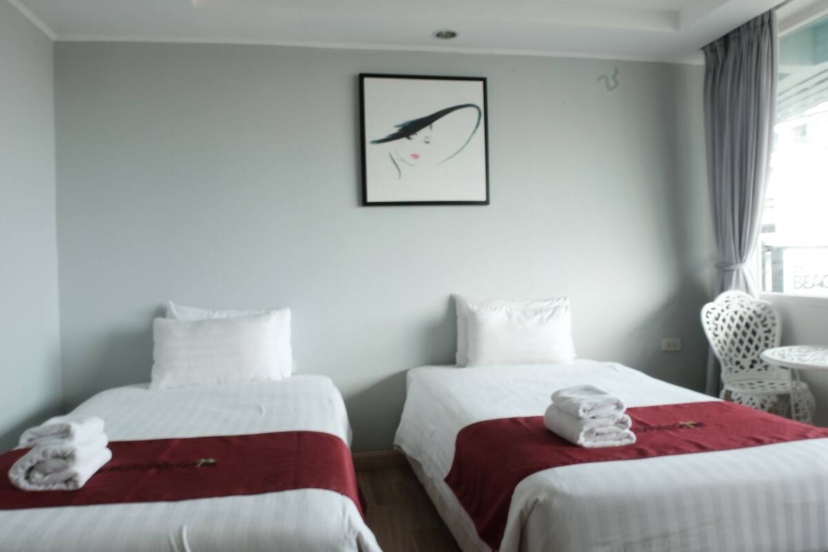 A hotel room with two beds, a red throw blanket, a white pillow, a framed artwork, and a white chair by the window.