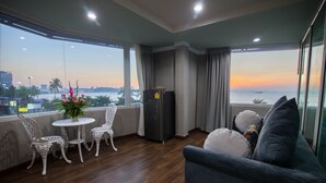 Un balcón con vistas a la puesta de sol sobre el agua, una pequeña mesa con sillas, un refrigerador y un sofá con cojines.