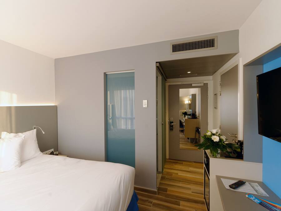 Habitación de hotel con cama, escritorio con televisor, espejo y puerta que da a un pasillo.
