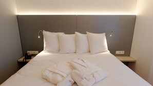 A neatly made bed with white bedding, a set of white slippers, and a robe on the bed. There are two bedside tables, one with a phone and the other with a lamp.