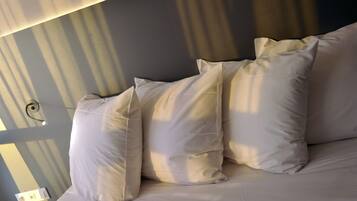 Una cama con ropa blanca y almohadas, un interruptor de pared y una lámpara en el techo.