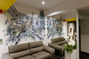 A room with a large mural of a cityscape on the wall, two brown sofas, and a white flower arrangement.