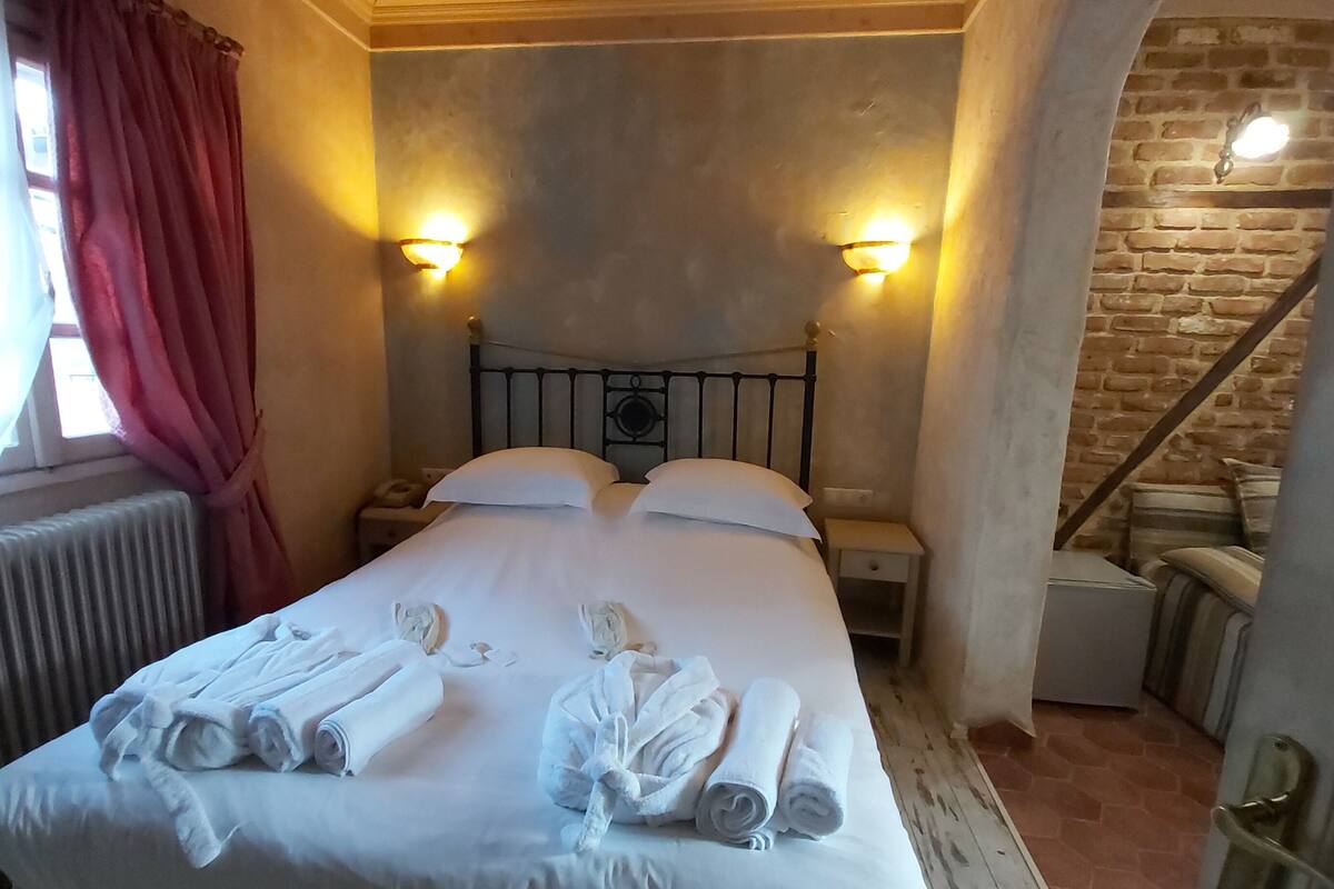 A bedroom with a bed, two bedside lamps, a radiator, a window with curtains, and a brick wall.