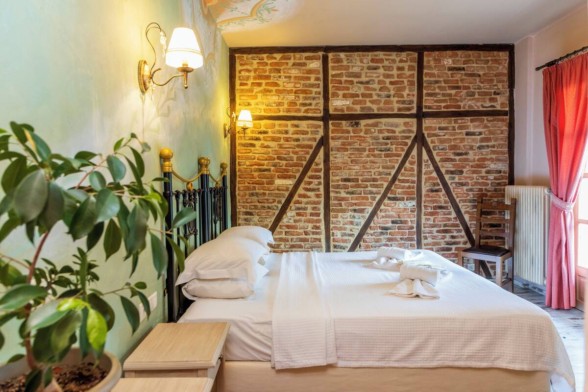 A hotel room with a brick wall, a bed with white linens, a potted plant, and wall-mounted lamps.