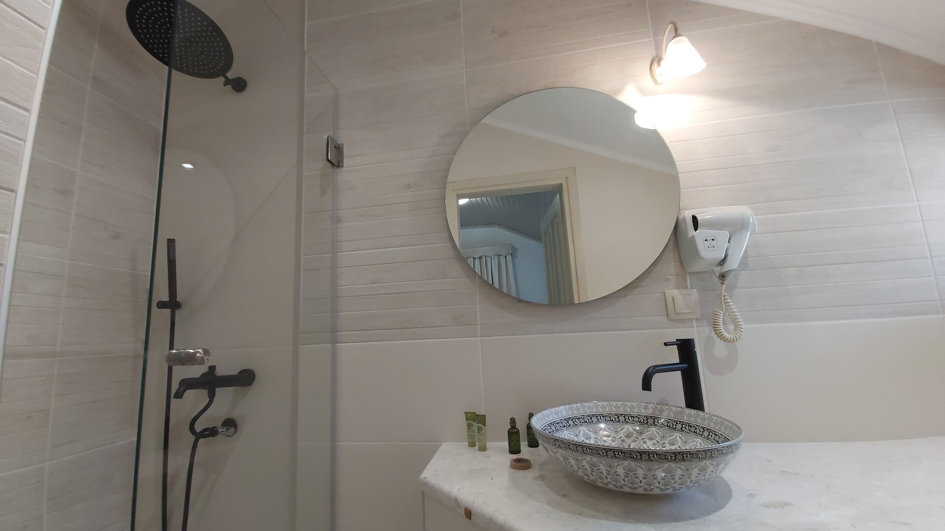 Un baño con ducha, un espejo redondo, un lavabo con un recipiente decorativo y un secador de pelo sujeto a la pared.
