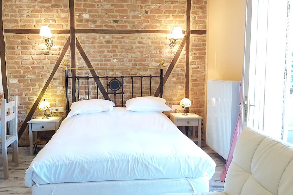 A bedroom with a brick wall, a bed with white bedding, two bedside tables with lamps, a wooden chair, and a window with curtains.