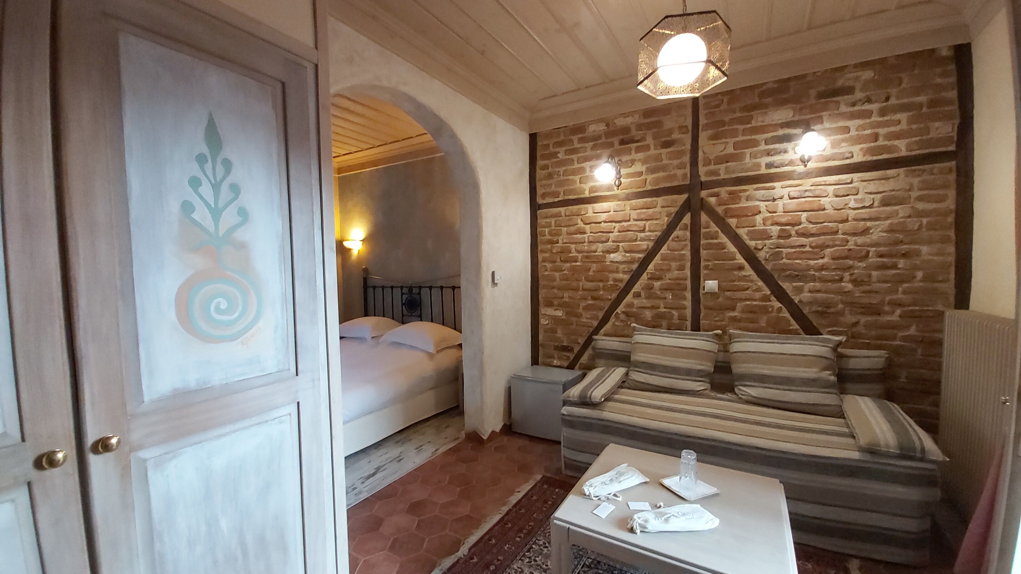 Una habitación con cama, sofá, mesa con objetos, pared de ladrillo y techo de madera.