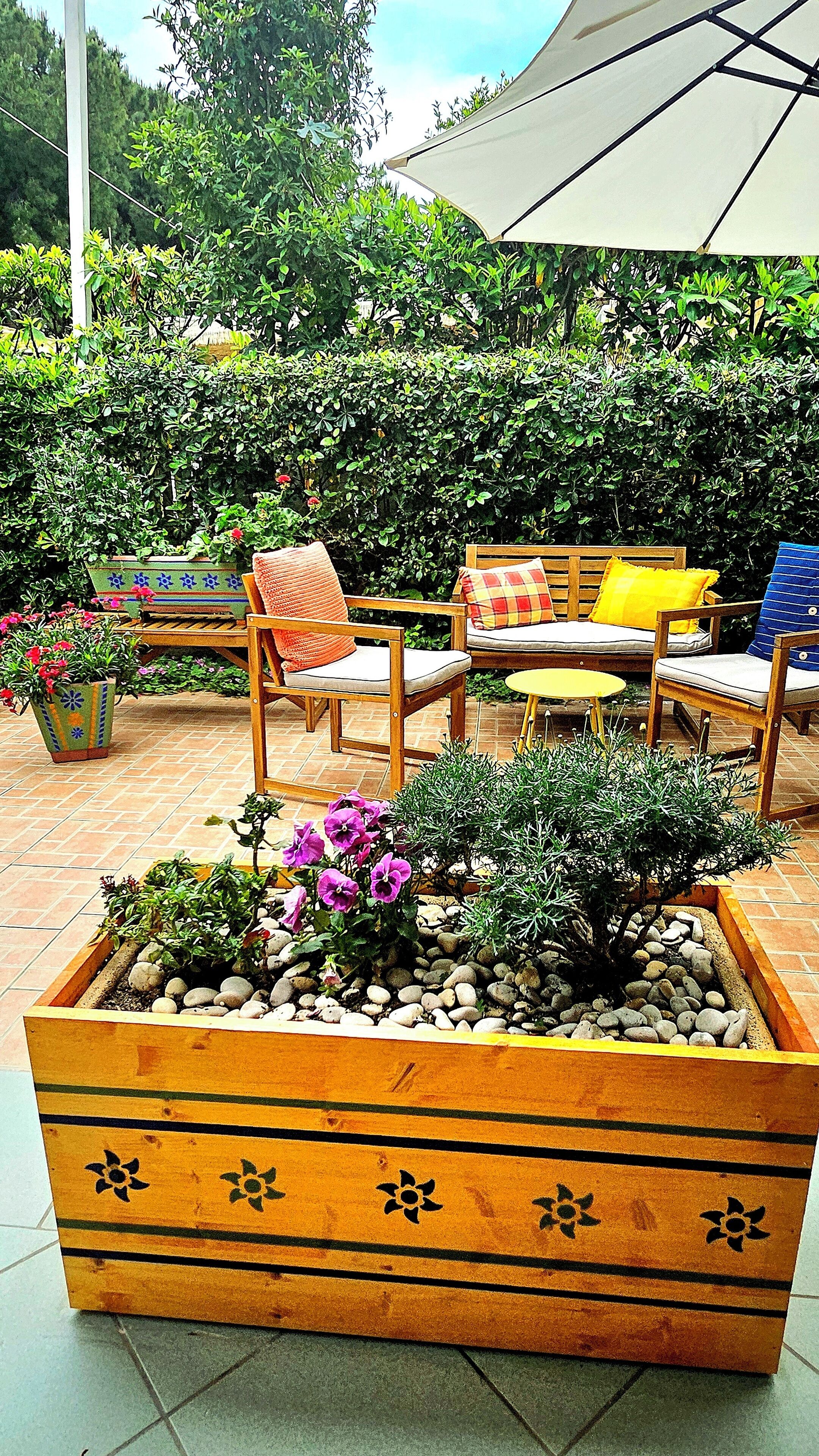 A wooden planter box with flowers and pebbles, surrounded by outdoor furniture including chairs and a table.
