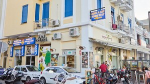A multi-story building with a blue and white facade, featuring various shops on the ground floor. - Amalia Rooms (Chios)