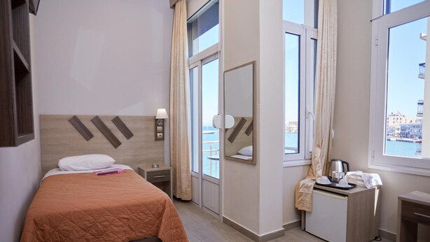 A hotel room with a bed, a nightstand, a desk with a telephone, a mirror, and a window with a view of buildings. - Amalia Rooms (Chios)