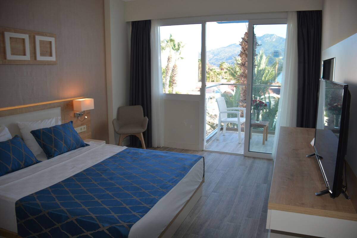 Une chambre d’hôtel équipée d’un lit, d’un bureau, d’une chaise, d’une télévision et d ’un balcon offrant une vue sur les palmiers et les montagnes.
