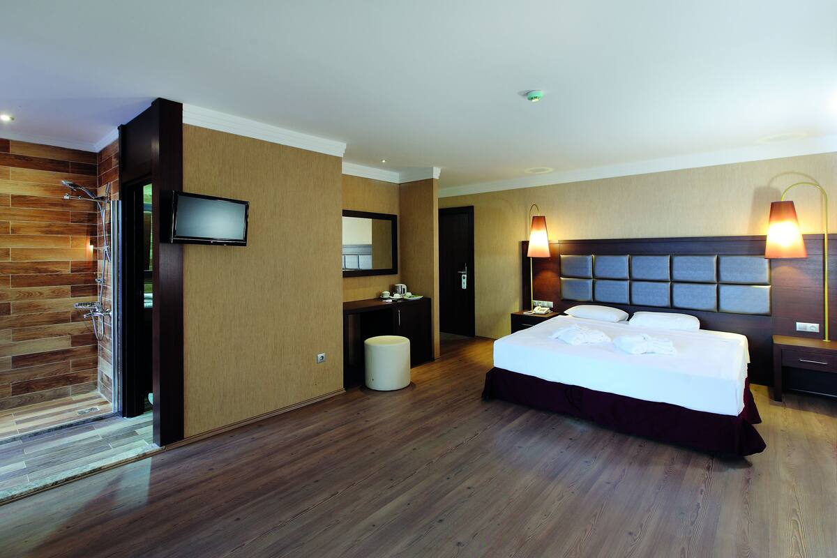 Une chambre d’hôtel avec un grand lit, un téléviseur fixé au mur, un plancher en bois et une salle de bain avec douche et toilettes.