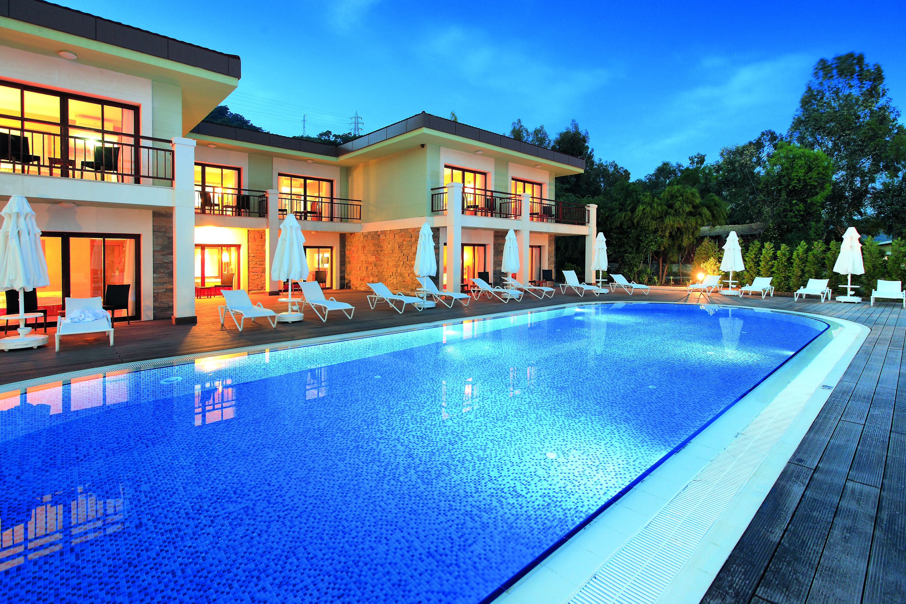A swimming pool with lounge chairs and umbrellas, a multi-story building with balconies, and a clear sky.