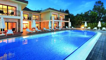 A swimming pool with lounge chairs and umbrellas, a multi-story building with balconies, and a clear sky.
