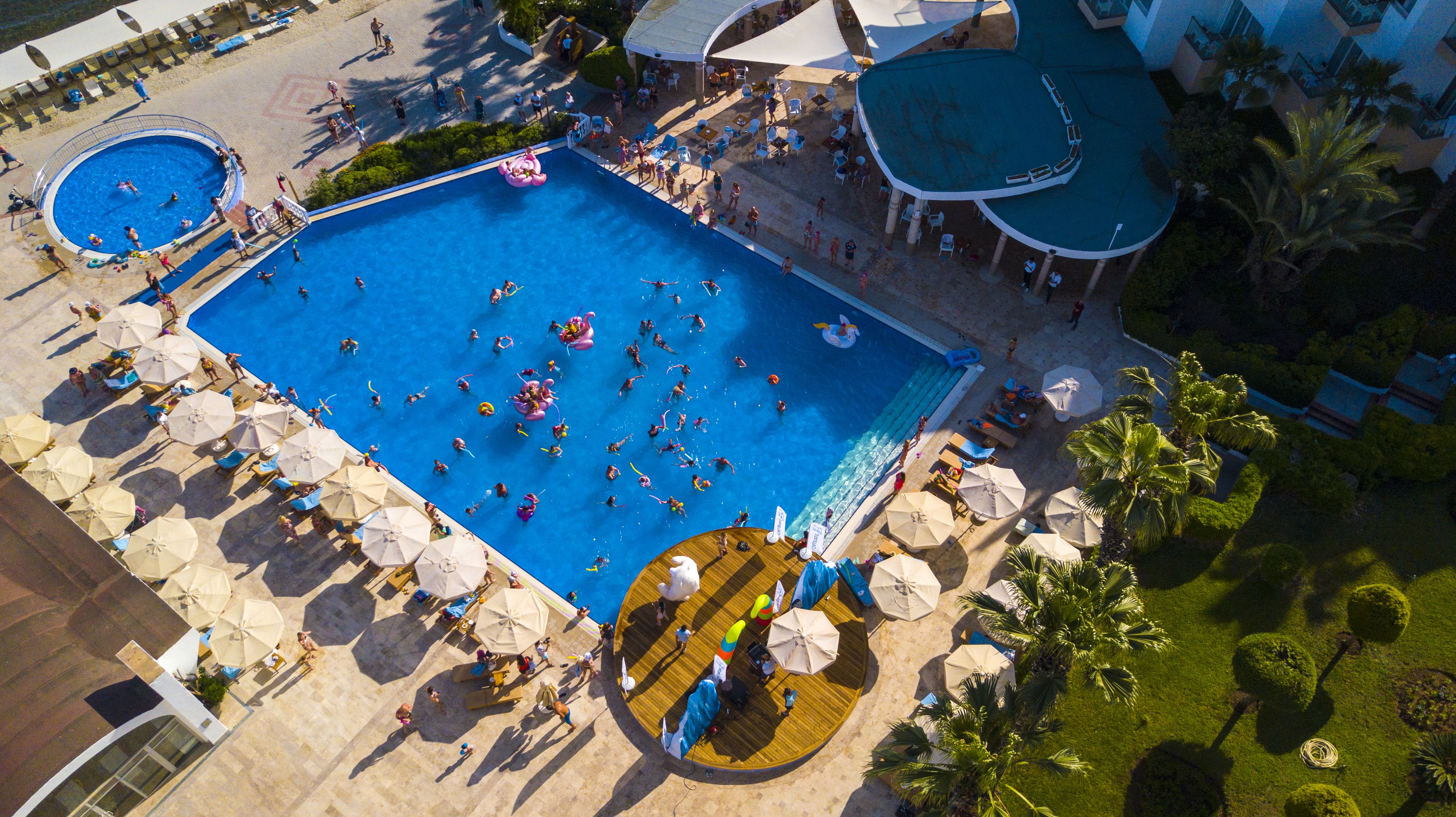 An aerial view of a resort with a large central pool surrounded by sun loungers and umbrellas, a smaller pool, and a covered area with seating.