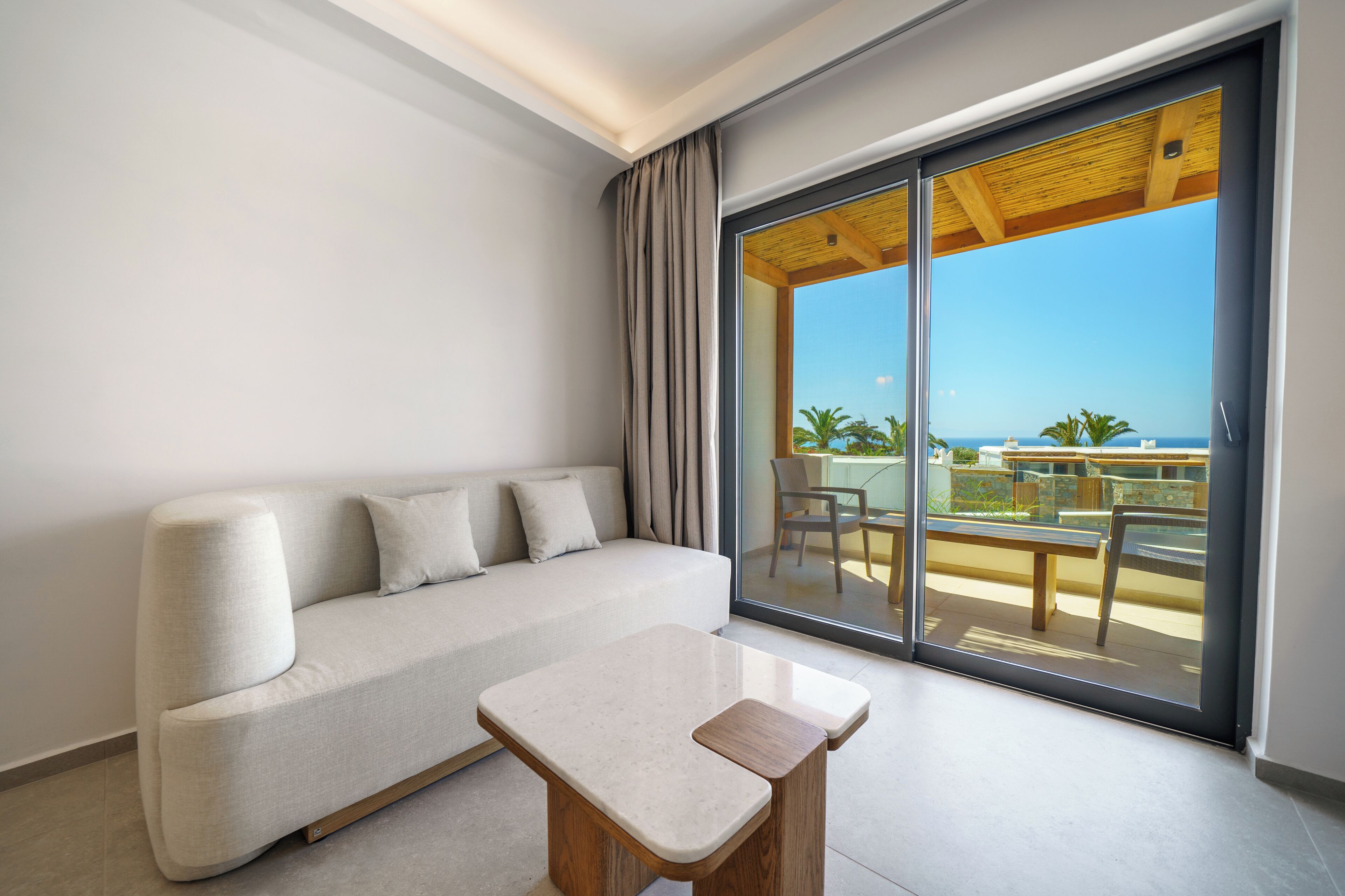 A modern room with a sofa and a small table, a sliding glass door leading to an outdoor area with a view of the sea.