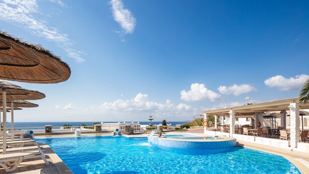 A swimming pool with lounge chairs and umbrellas, a covered seating area, and a view of the ocean. - Kavuras Village Hotel & Suites (Naxos)