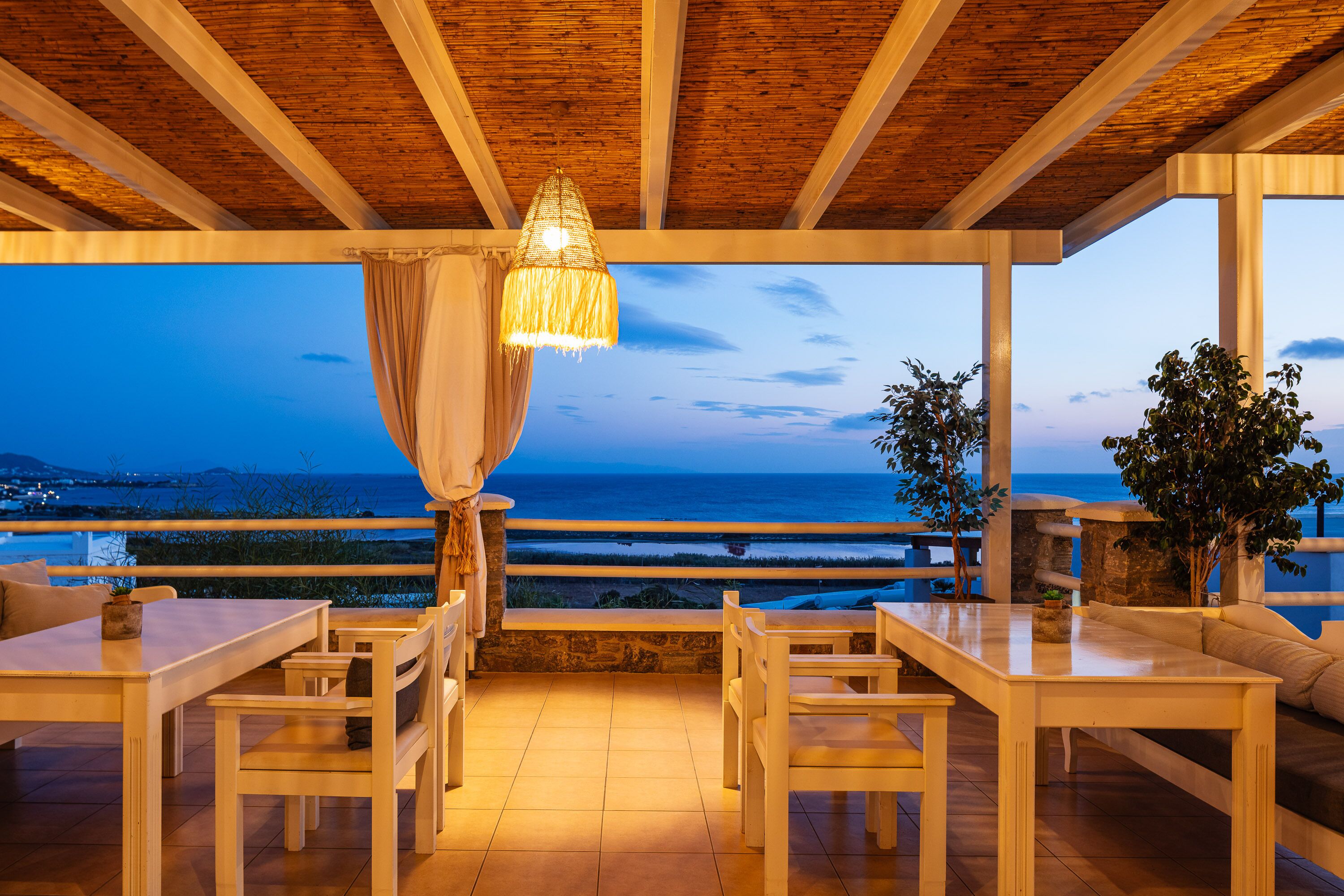 Un área de comedor al aire libre con mesas y sillas de madera, una lámpara colgante y vista al océano al atardecer.