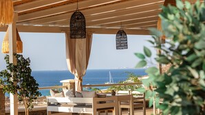 An outdoor seating area with a view of the sea, featuring a wooden table, chairs, and hanging wicker lamps. - Kavuras Village Hotel & Suites (Naxos)