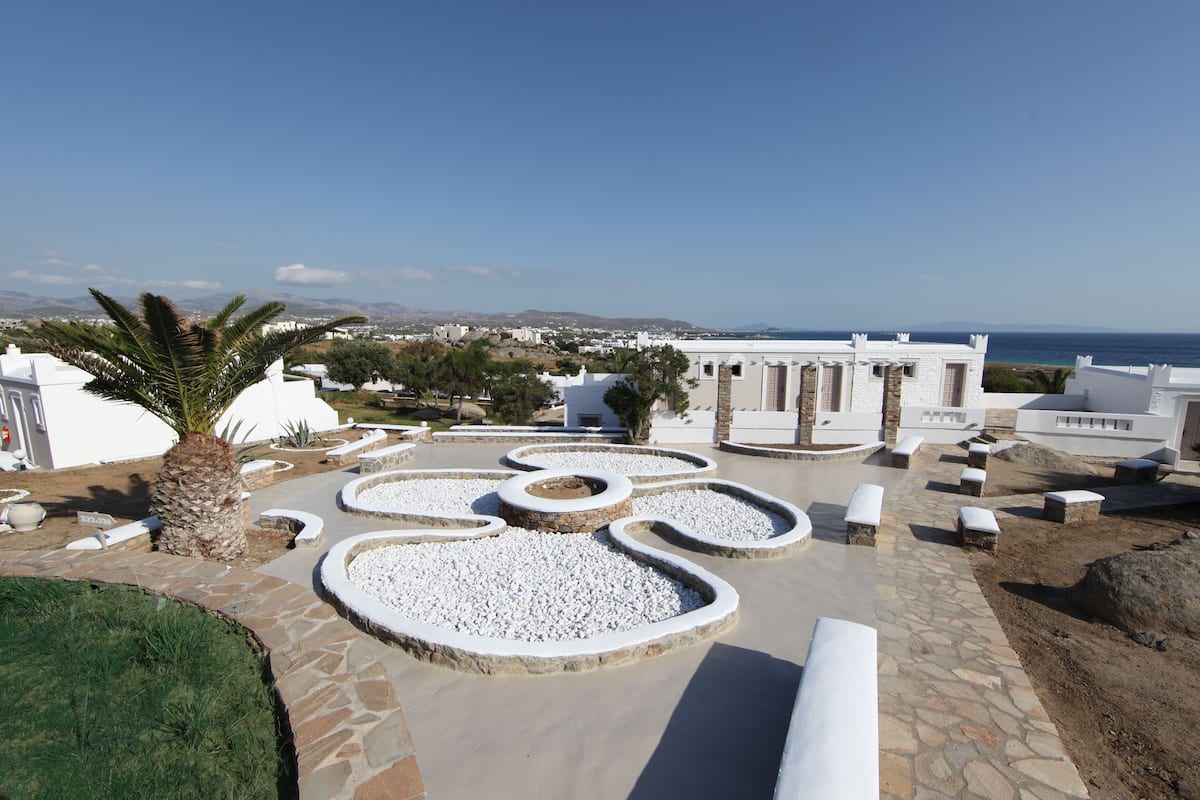 A landscaped courtyard with white stone planters, a palm tree, and a view of the sea and distant buildings.