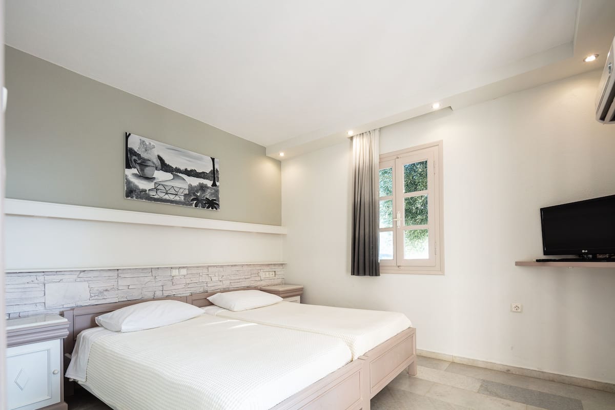 A bedroom with a bed, a nightstand, a TV, a window with curtains, and a painting on the wall.