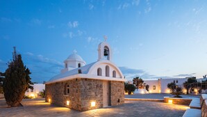 A small stone church with a bell tower and two domes, surrounded by a paved courtyard with stone benches and a large tree. - Kavuras Village Hotel & Suites (Naxos)